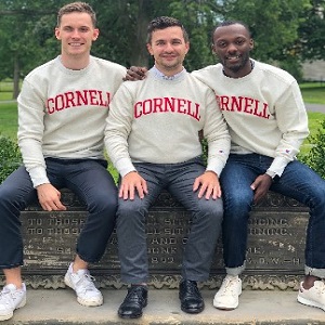 Image of three EMBA Metro NY students sitting on a bench outside, wearing Cornell University sweatshirts