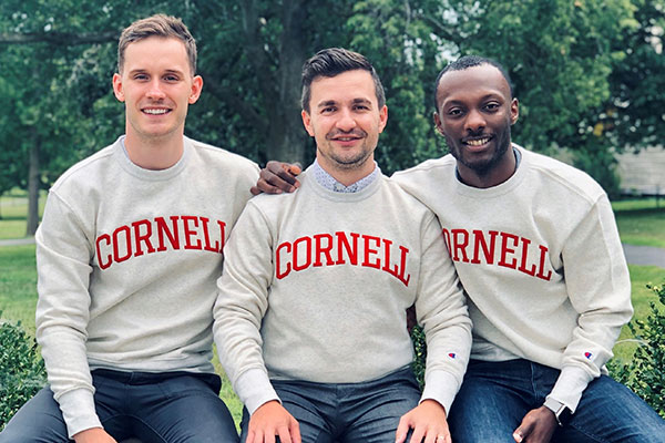 Image of three EMBA Metro NY students sitting on a bench outside, wearing Cornell University sweatshirts