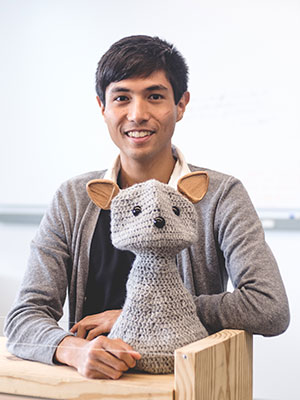 Michael Suguitan sits at a desk behind a crocheted social robot he helped create. 