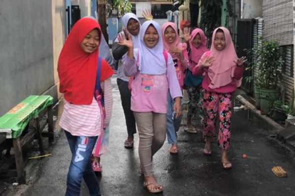 A group of smiley young Indonesian girls wearing pants and hajibs are waving as they walk between buildings