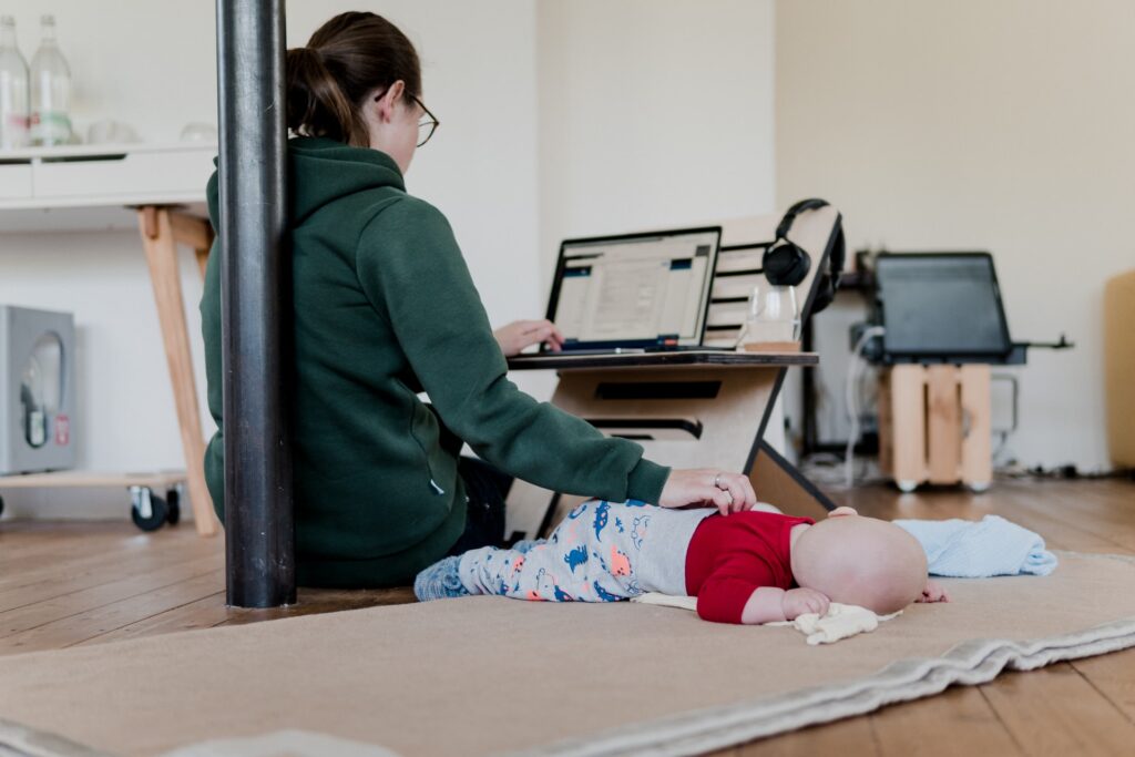 A woman sooths a baby while doing her best to also work on her computer. Caption: For women especially, working from home may come with the added pressure to juggle more responsibility.