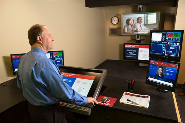 Andrew Karolyi in the studio at Sage Hall teaching students in the Executive MBA Americas program via real-time videoconferencing. He is standing and has views of students in different cities on different screens and other screens with information too