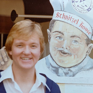 1981 photo of Andrew Karolyi standing in front of a sign depicting a chef in a chef's hat with "Schnitzel House written on it