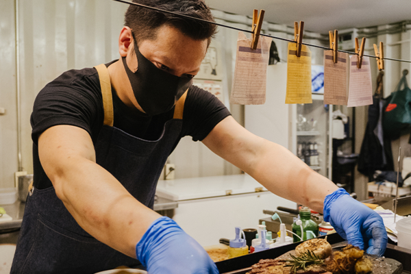 A chef working in a kitchen with a mask and gloves on, setting a plate of food onto a tray.