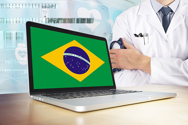photo of laptop with the Brazilian flag on the screen and in the background, just the right shoulder and left hand of a medical professional in a white lab coat, shirt and tie, holding a stehoscope