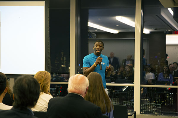 photo of Mousa Paye standing in front of an audience and speaking. Behind him is a window that is reflecting the audience (it is night out)