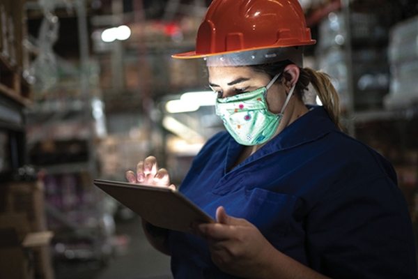 Person wearing a face mask and hardhat works on a touch screen in a warehouse.