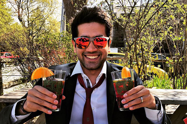 ankit Dhawan sitting outside, grinning, wearing sunglasses, in warm weather and holding up two glasses of a beverage with orange slices on the rims