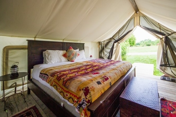 Inside a typical Firelight Camps tent featuring a large bed with a colorful bedspread, wood floor, chest, and table and a camping lantern.