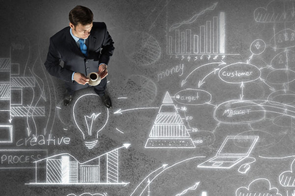 Business person superimposed onto chalkboard-like background with idea bubbles, graphs, charts, technologies, and flow charts, all meant to depict business strategizing.