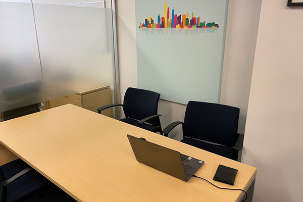 A picture of a small meeting room with a glass wall, a desk, two chairs, and a laptop.