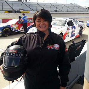 Gage Javier standing and hold a helmet at a race track with race cars in the background