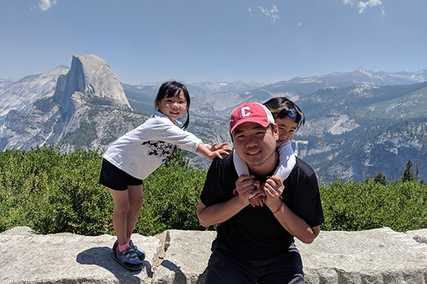Chao Yang with one small girl climbing on his back and another leaning on his shoulder with green and rocky mountainous scenery and blue sky in the background