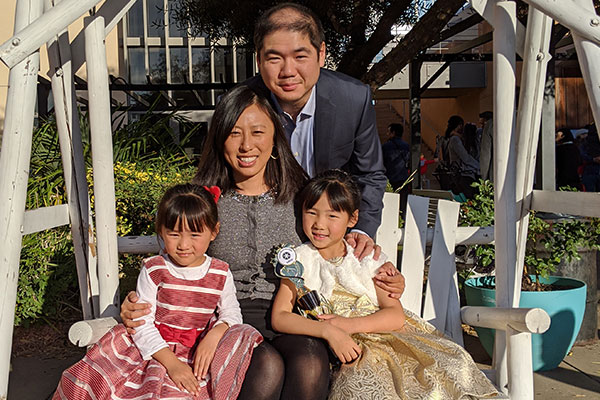photo of Chao Wang and Elain Qin sitting on a bench swing with their young daughters, smiling and all dressed up