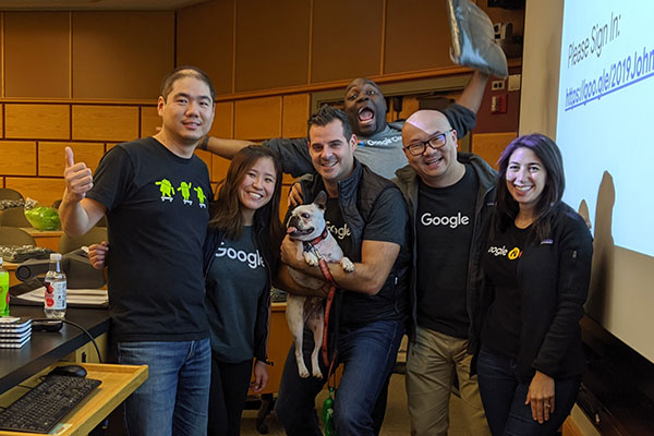Chao Wang with 5 other people, all wearing Google T-shirts and smiling, at the front of a lecture hall. One is holding a small dog and one is jumping up in the background 