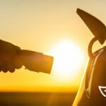 photo of a hand holding an electric charger and about to insert it into the side of a car with a burst of light from the rising sun in the gap between the charger and the car