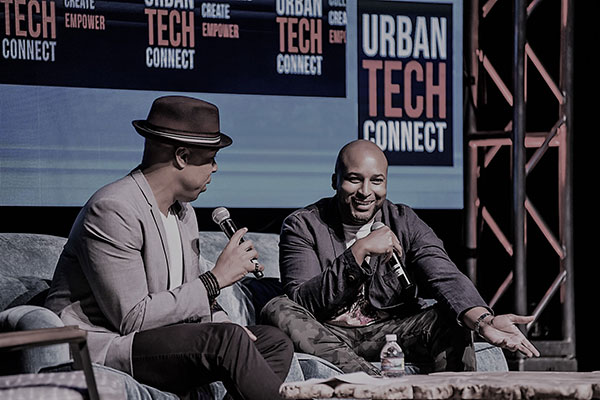 Marlon Nichols sitting on a couch with one other man, both holding microphones, with Urban Tech Connect signs displayed on the wall behind them