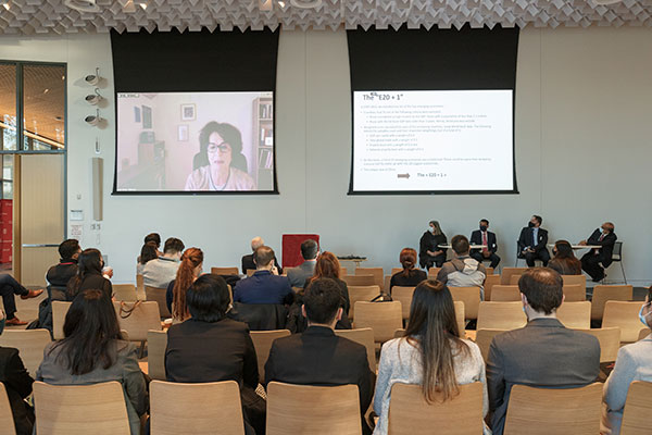 photos of people sitting chairs in the foreground, looking at two screen on the wall at the front of a lecture hall. Ann Miroux is on one screen and the other is a slide about the E20