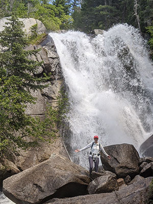 photo of waterfalls and a tiny image of Julie Belkus climbing among big boulders situated across from the waterfalls