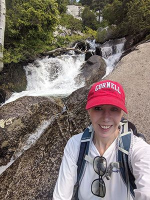 photo of Julie Belkus in the foreground, smiling, wearing a backpack and red Cornell hat, beside a waterfalls