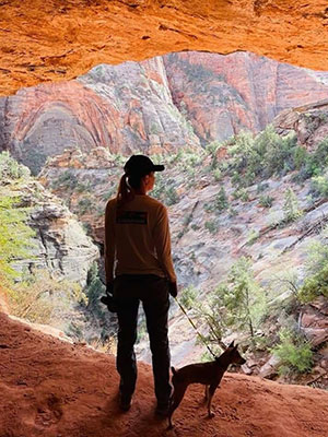 photo of Julie Belkus and her small dog, her back to the camera, overlooking a deep canyon with red rock above and some vegetation in the canyon