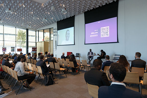 photo of people sitting in a lecture hall and looking at screens on the wall at the front of the room. Múyìwá Mátùlúkò is on one of the screens.