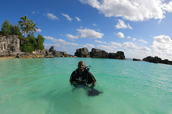 Pradeep Ambrose standing up in aqua blue water with the beach and trees and rocks in the distance, blue sky abovein scuba diving gear 