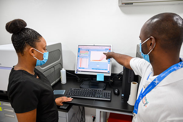 Pradeep Ambrose points to a screen with graphs while a women on his left looks on (both are wearing face masks)