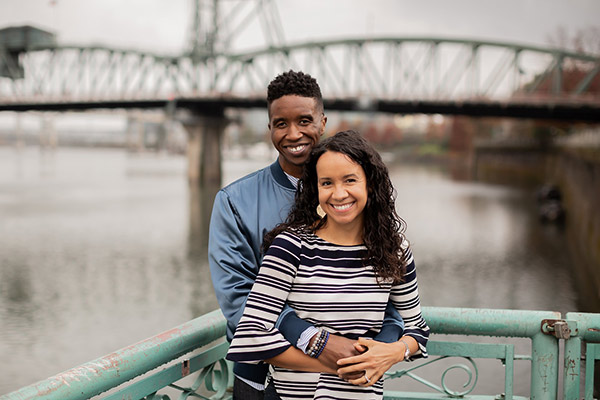 Aaron Seabron standing with his arms around his wife, who is standing in front of him, with a river and bridge in the background