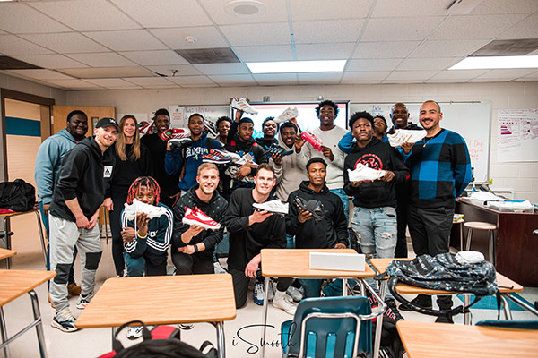 A group of group of 17 boys and men and one woman standing or half kneeling in a classroom, smiling and holding up Adidas shoes