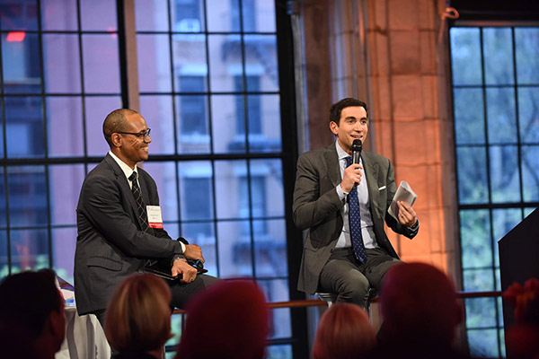 Morgan Jones and Andrew Sorkin seated on stools and speaking into microphones on a stage with a big window in the background and the audience, seated, in the foreground