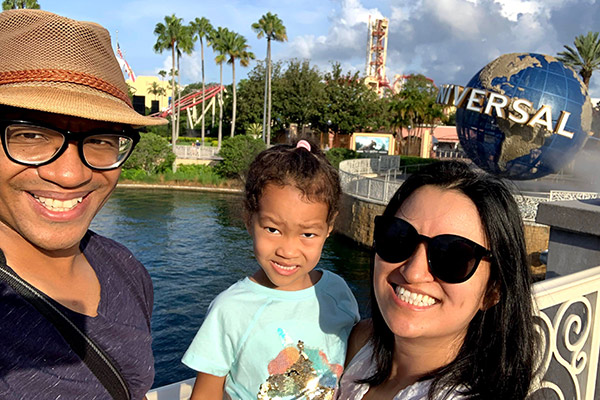 Morgan Jones standing next to his wife, who is holding their young daughter, with a pond, palm trees, green grass, and a Universal Studios globe in the background