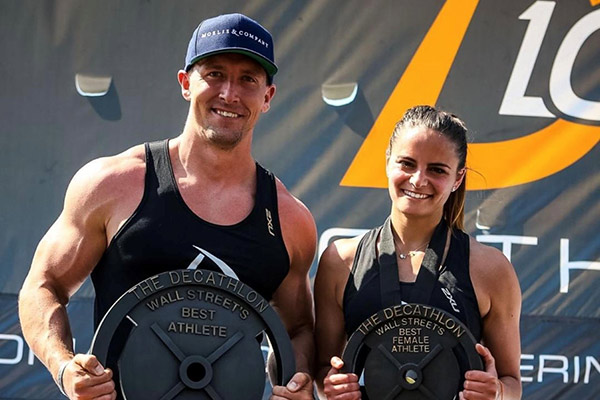 Thomas White stands next to a woman, both in athletic gear and both holding trophies that say "Decathlon, Wall Street, Best Athlete" and "Decathlon, Wall Street, Best Female Athlete"