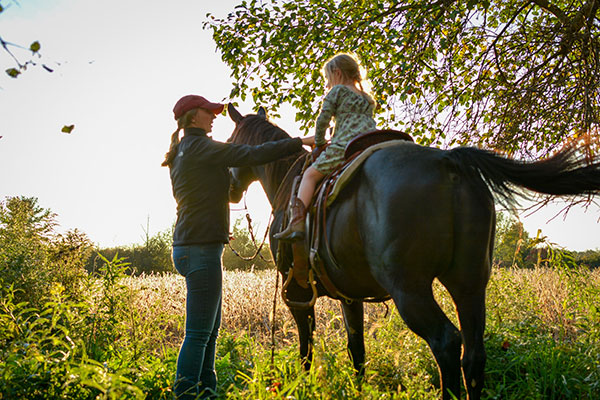 Anna Richards standing next to and holding her horse's reins with her young daughter sitting in the saddle. They are in a green field, in the shade under a tree