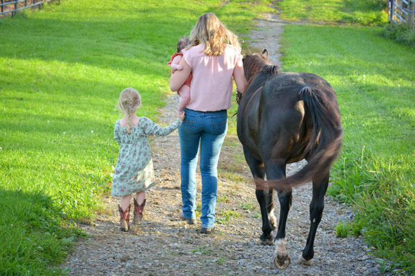Anna Richards walking away from the camera side by side with her 3-year-old daughter and horse and carrying her infant