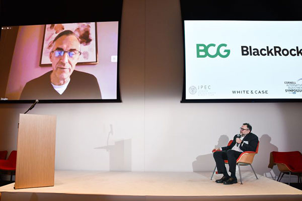 1 man seated in a chair on stage and 1 man on screen talking to one another