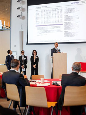 3 men and one woman, one of the men standing and speaking at a podium with a table and symposium attendees in the foreground