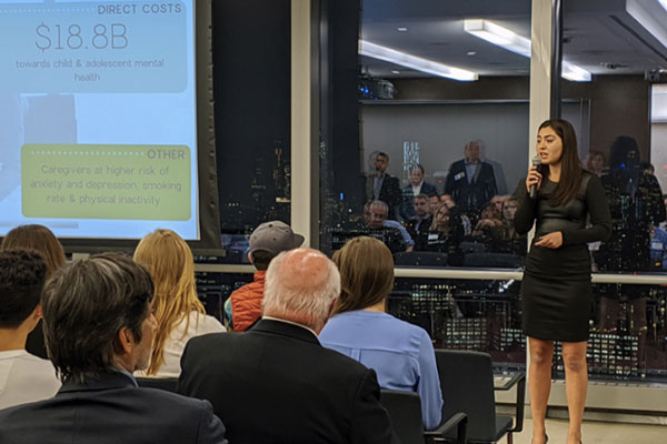 Marla Beyer presenting to a room full of people sitting in chairs, dark window with their reflection behind her and further to the left, a screen