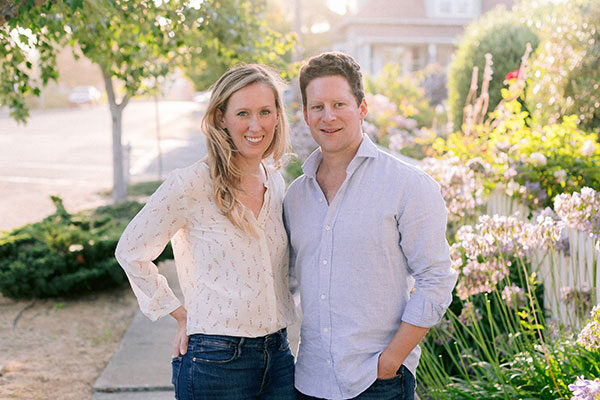 Julia and Alex Littauer stand outside on a city street with foliage in the background sunny, spring day.