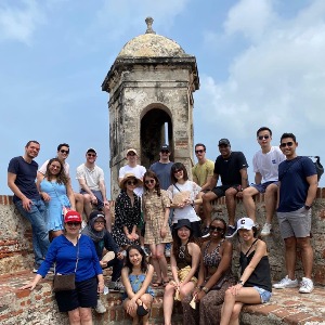 a group of about 20 students surround the tops of an old-looking tower with blue sky in the background.