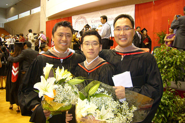 three young Korean men in graduation robes and hoods holding huge bouquet of lilies with a stage and other people in the backround