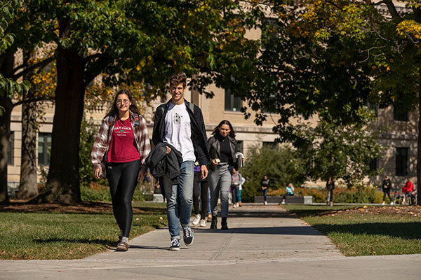 Students walking on sidewalk paths under a canopy of trees with a college building in the background.