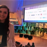 A picture of Ina Gjika at the opening plenary of the conference with the stage and big screen of sponsors behind her.