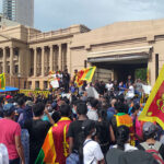 A large crowd of people, some waving Sri Lanka’s flag, facing government buildings and listening to speakers standing in front of those buildings.