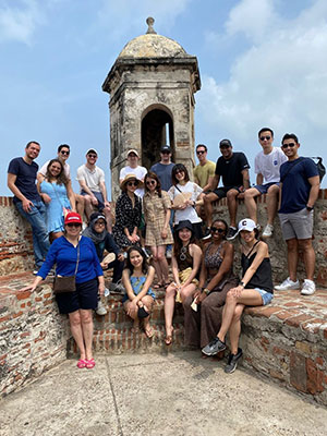 A group of about 20 students seated and standing on stone walls surrounding a stone tower with sky in the background.
