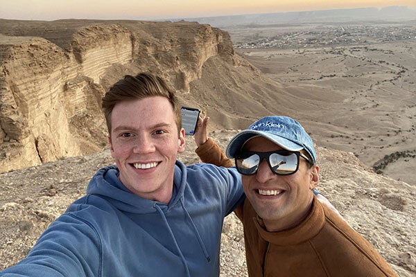 Doug Barnard and a young Saudi friend take a selfie with a vast sandy dessert and sand dunes in the background.