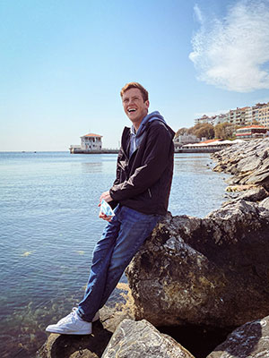 Doug Barnard standing on a rocky shoreline with more water and buildings in the background.