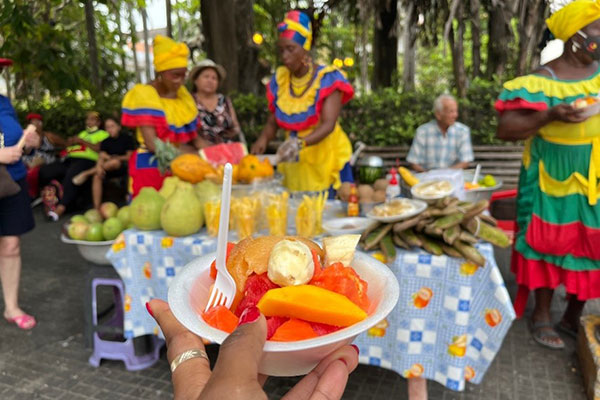 in the foreground, a hand holding a colorful bowl of fruit. In the background, woman servers dressed in colorful attire surround a table laden with many different kinds of fruit. People sit on benches in the background.