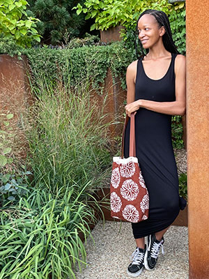 A young Black woman in a black tank dress and sneakers standing and leaning against a wall in a garden surrounded by green foliage, holding a brown and white print tote bag.
