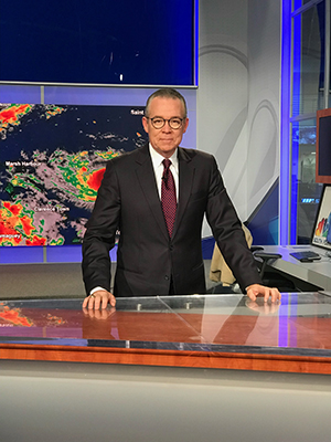 John Morales behind a news desk in a TV studio with a weather map behind him.